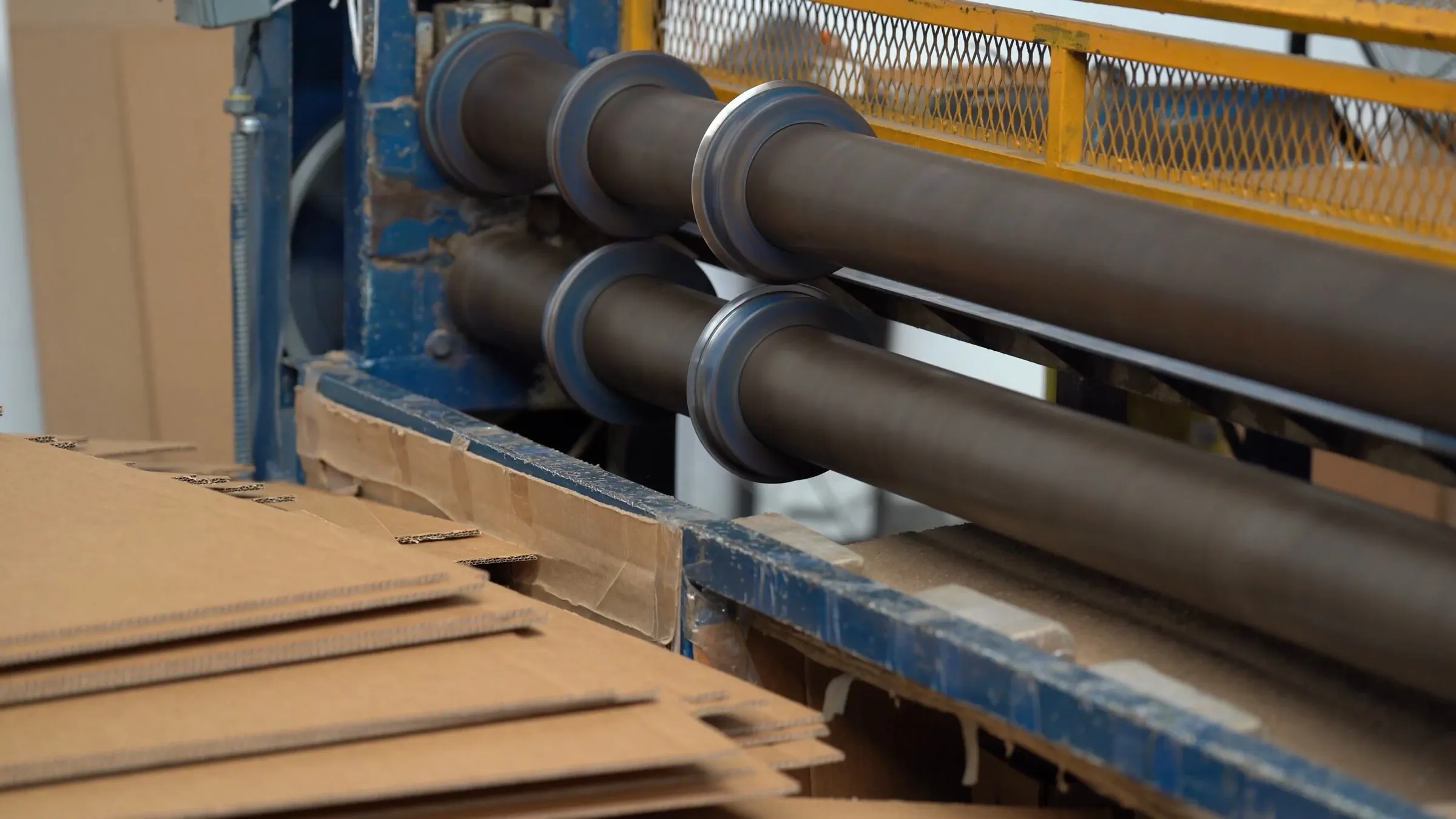 Brown corrugated sheets stacked beside the rollers of a slitting and scoring machine in a box plant.