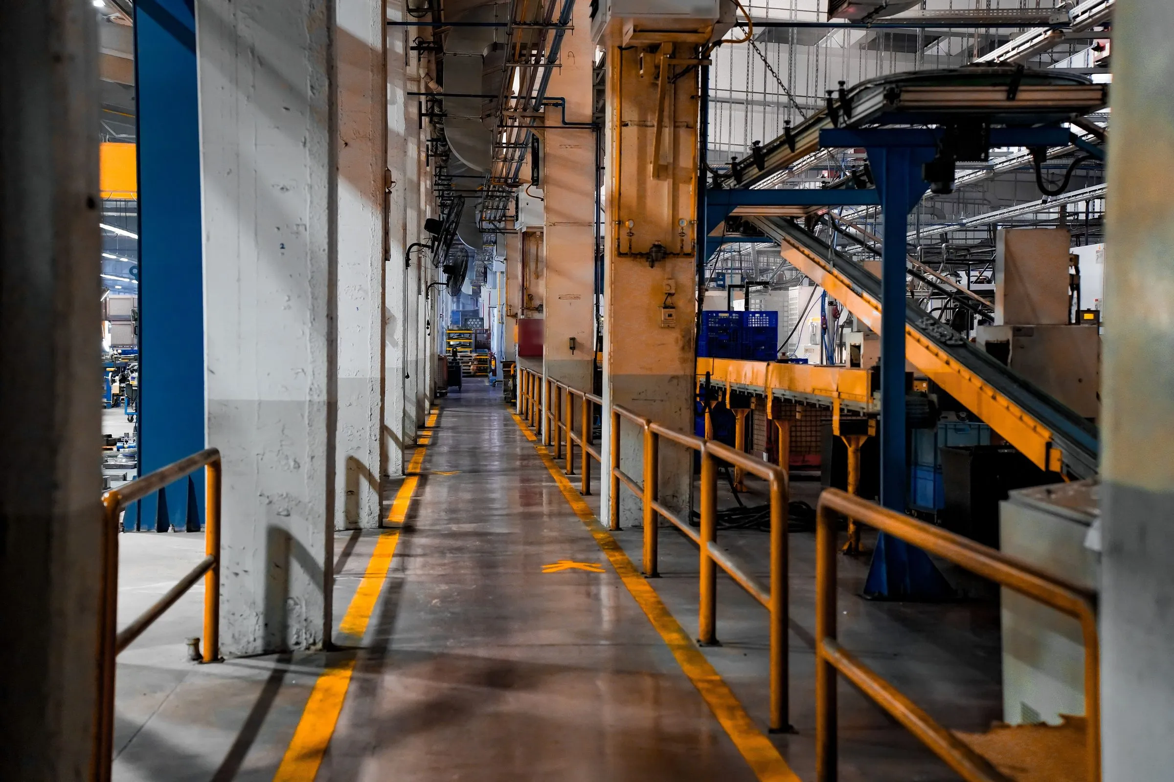 Long walkway with yellow safety lines and railings running beside conveyors inside a factory.